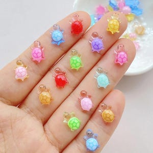 Puede incluir: Una colección de pequeños abalorios con forma de tortuga de colores. Los abalorios translúcidos vienen en varios colores, incluyendo rojo, azul, verde, amarillo, rosa y morado. Estos abalorios decorativos son ideales para manualidades y bisutería.