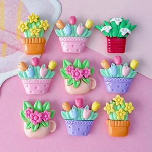 May include: A collection of colorful, miniature flower pot and cup decorations. The assortment includes pots and cups in orange, pink, and purple, each filled with vibrant flowers in shades of yellow, pink, and white. The background is a soft pink.
