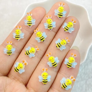 Puede incluir: Una colección de pequeños adornos con forma de abeja, amarillos y negros. Cada abeja tiene una cara sonriente, antenas naranjas y alas azules translúcidas. Las abejas están dispuestas sobre una superficie clara.