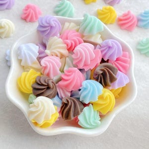May include: A white ceramic bowl filled with colorful, swirl-shaped resin embellishments. The embellishments are in various pastel colors, including pink, yellow, blue, green, and purple, and a few are brown. These decorative pieces resemble miniature frosting swirls.