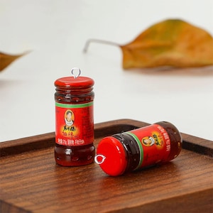 May include: Two miniature red and brown sauce bottles with silver loops on top, one upright and one on its side. The bottles have red labels with Chinese text and a green stripe. They sit on a wooden tray.