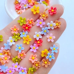 Peut inclure: Une collection de petites décorations en forme de fleurs multicolores, dans des tons de jaune, orange, rose, rouge, bleu et vert. Chaque fleur a un centre jaune. Les fleurs sont dispersées sur un fond clair.