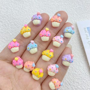 May include: A collection of miniature cupcake charms in various colors, including pink, yellow, blue, and purple. Each cupcake has a white base and is topped with colorful frosting and a heart-shaped decoration. The cupcakes are small, likely for crafting or jewelry making.