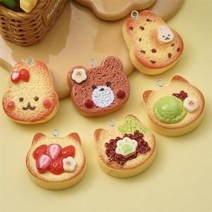 May include: Six colorful resin charms shaped like toast with various toppings, including a bear, a cat, a strawberry, and a green swirl. Each charm has a silver loop for attaching to a necklace, bracelet, or keychain.