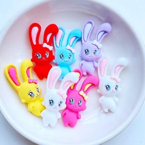 以下が含まれることがあります： 白いボウルに入った、カラフルなウサギ型の装飾品のコレクション。ウサギは赤、黄、青、紫、白、ピンクなど様々な色があります。各ウサギにはリボンと詳細な顔の特徴があります。