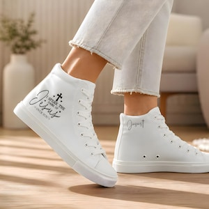 Könnte beinhalten: Weiße High-Top-Sneaker mit schwarzem Text "Speak the name of Jesus over you" und einer Kreuzgrafik. Das Wort "Jesus" ist auch auf der Seite des anderen Schuhs aufgedruckt. Die Sneaker haben weiße Schnürsenkel und Sohlen.