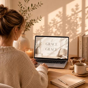 K&ouml;nnte beinhalten: Eine Person arbeitet an einem Laptop auf einem Holztisch. Der Laptop-Bildschirm zeigt die Worte "GRACE UPON GRACE". Eine brennende Kerze, eine Tasse Kaffee, ein Notizbuch und B&uuml;cher befinden sich ebenfalls auf dem Schreibtisch. Eine Vase mit Gr&uuml;nzeug ist im Hintergrund.