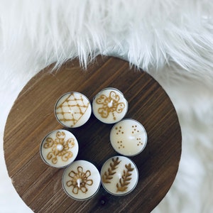 May include: Six white tea light candles with gold designs on a wooden surface. The designs include a diamond, a flower, a leaf, and dots.