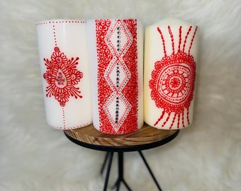Puede incluir: Tres velas blancas con diseños de henna rojos. Las velas están sobre un soporte de madera con patas de metal negro.