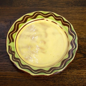 May include: A round ceramic plate featuring a smooth, pale yellow center and a wide, light green rim. The rim is decorated with a continuous, dark brown wavy line pattern that follows the circular edge of the plate, creating a distinct contrast.