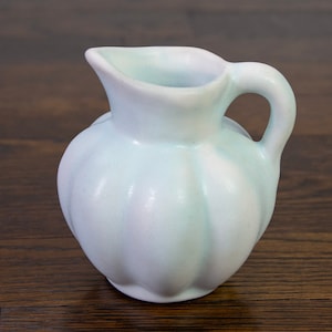 Puede incluir: Jarra pequeña de cerámica azul claro, cuerpo redondeado y asa. Presenta un pico vertedor y una textura acanalada. Diseño sencillo y elegante, ideal para servir líquidos. Mide aproximadamente 15 cm de altura.