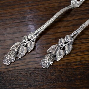 May include: Two silver serving utensils with floral rose and leaf designs. The handles have a textured pattern. The utensils are on a dark wood surface.