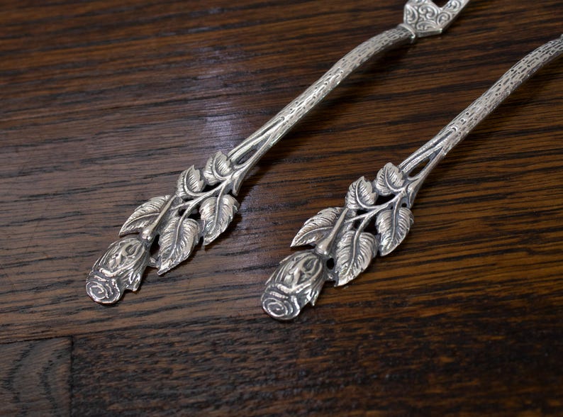 May include: Two silver serving utensils with floral rose and leaf designs. The handles have a textured pattern. The utensils are on a dark wood surface.