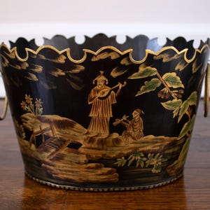 May include: A black metal bucket with a scalloped top edge and two gold-toned ring handles. The exterior features hand-painted gold and green illustrations of figures playing musical instruments, trees, and a bridge in a landscape setting.