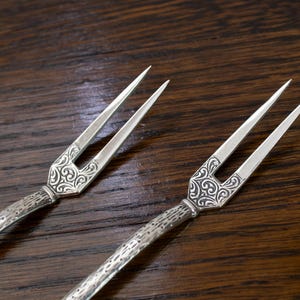 May include: Two silver serving forks with ornate, engraved handles and two tines each. The forks are displayed on a dark brown wooden surface, showcasing their intricate design and elegant appearance.