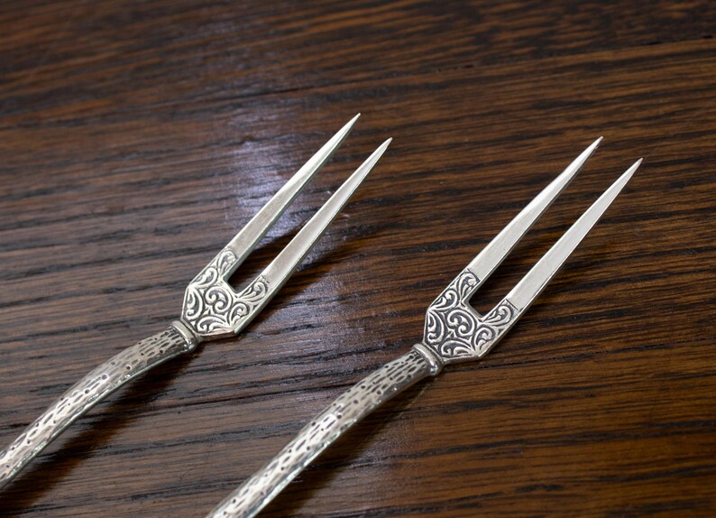 May include: Two silver serving forks with ornate, engraved handles and two tines each. The forks are displayed on a dark brown wooden surface, showcasing their intricate design and elegant appearance.