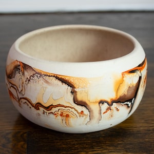 Puede incluir: Un cuenco redondo de cerámica blanquecina con una amplia abertura. El exterior presenta patrones abstractos en tonos marrones, naranjas y crema. El cuenco se encuentra sobre una superficie de madera oscura, mostrando su diseño decorativo.