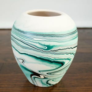 May include: A white and teal marbled ceramic vase with a rounded shape. The vase features a swirling pattern of teal, black, and white, creating a unique, artistic design. The opening is a light beige color.