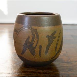 May include: A brown ceramic vase with a dark brown glaze and abstract black brushstrokes. The vase has a rounded shape and a wide opening.