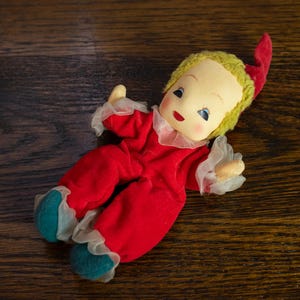 Puede incluir: Una muñeca vintage con un atuendo de terciopelo rojo, una peluca amarilla y un gorro puntiagudo rojo. La muñeca tiene una cara pintada con ojos azules y boca roja. Los pies de la muñeca son turquesa con ribete de encaje blanco.