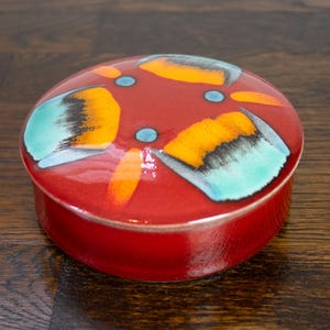May include: A round ceramic lidded box with a glossy red glaze. The lid features a decorative pattern of four stylized petal shapes in shades of orange, yellow, and turquoise, accented by small light blue dots arranged in a central cross formation.