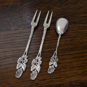 May include: Three antique silver serving utensils: two forks and a spoon. The forks have two tines and ornate handles with a leaf and floral design. The spoon has a rounded bowl and a similar leaf and floral handle design.