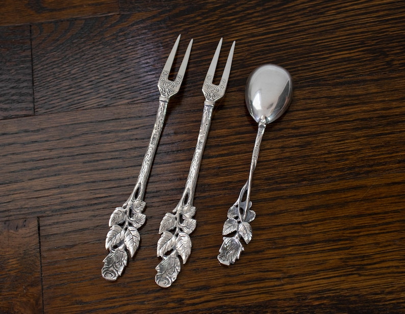 May include: Three antique silver serving utensils: two forks and a spoon. The forks have two tines and ornate handles with a leaf and floral design. The spoon has a rounded bowl and a similar leaf and floral handle design.