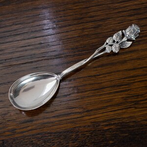 May include: A silver spoon with a rose design on the handle. The spoon has a polished, reflective bowl and a detailed rose and leaf motif. The spoon is likely made of sterling silver and is a decorative item.