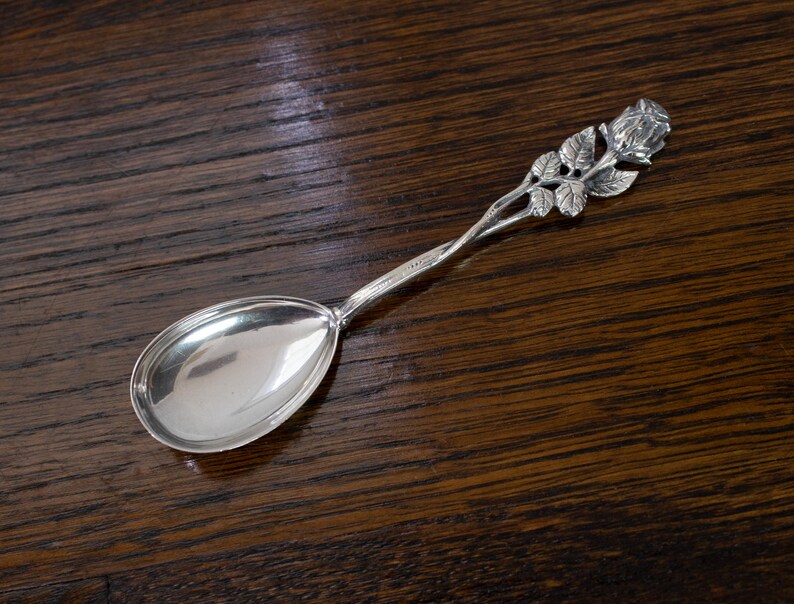May include: A silver spoon with a rose design on the handle. The spoon has a polished, reflective bowl and a detailed rose and leaf motif. The spoon is likely made of sterling silver and is a decorative item.