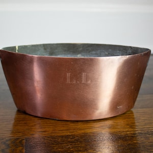 May include: A wide, shallow copper bowl with a metallic finish and a smooth, rounded rim. The exterior surface features the engraved letters L.L. centered on the front. The interior of the bowl has a dull, greyish metallic lining.
