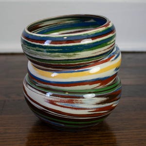 May include: A stack of three colorful ceramic bangles. Each bangle features a unique pattern of horizontal stripes in shades of blue, green, red, yellow, and white. The bangles are stacked on top of each other, showcasing their vibrant designs.