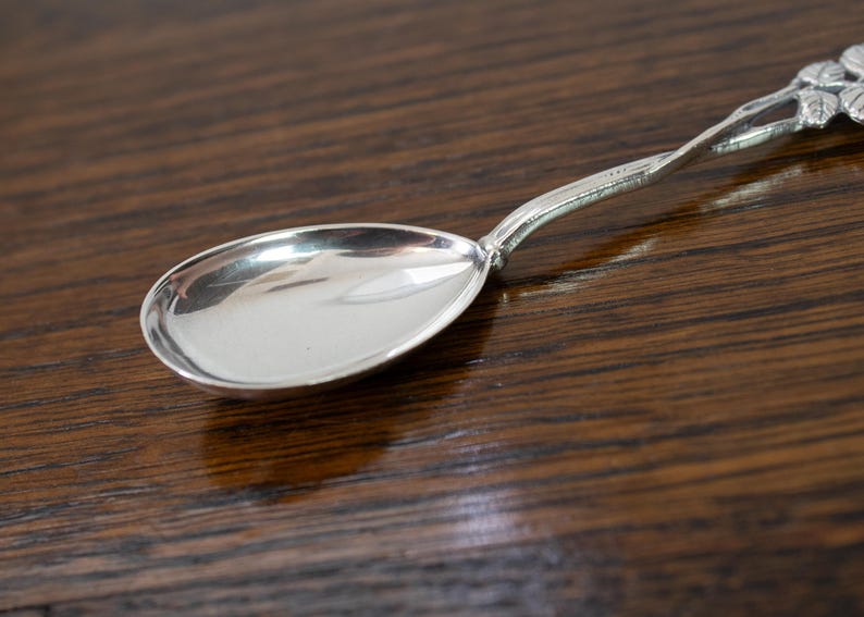 May include: A silver spoon with an oval bowl and a decorative handle. The handle features a floral design at the end. The spoon is set against a dark wood surface, highlighting its polished finish. This spoon is ideal for serving or eating.