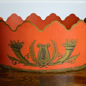 May include: A round orange metal planter with a scalloped top edge and gold trim. The exterior features a central gold lyre graphic flanked by two cornucopias. Two gold lion head handles with rings are attached to the sides of the container.