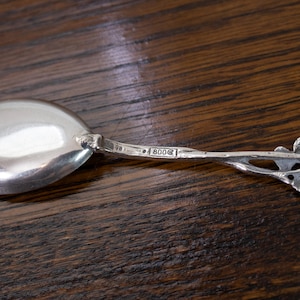 May include: A silver spoon with a rounded bowl and a handle featuring a leaf design. The spoon has the marking "800" on the handle. The spoon is set against a dark wood surface.