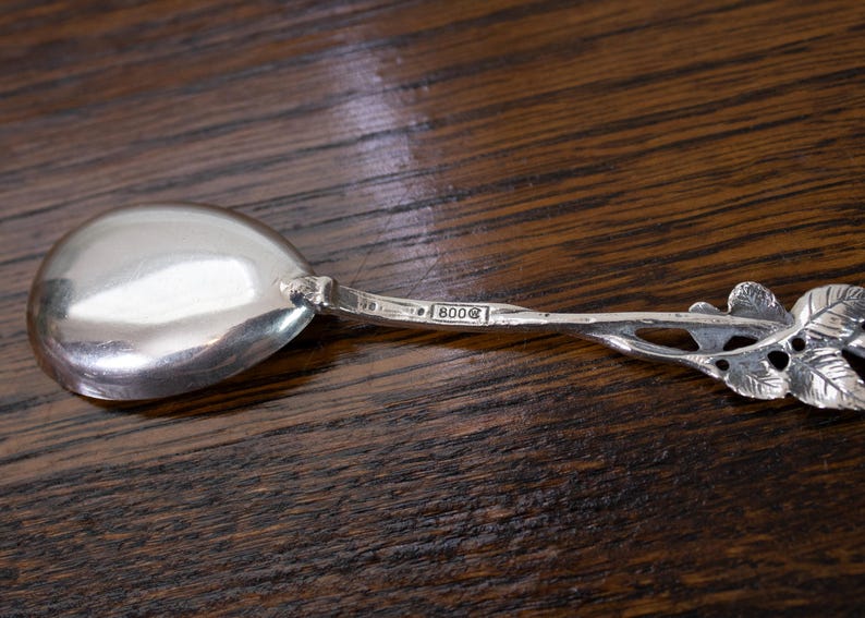 May include: A silver spoon with a rounded bowl and a handle featuring a leaf design. The spoon has the marking "800" on the handle. The spoon is set against a dark wood surface.
