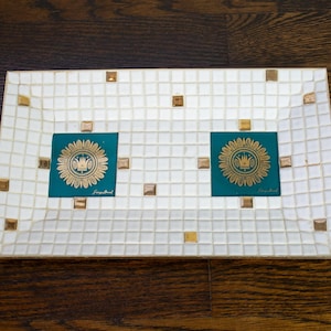 Puede incluir: Bandeja decorativa rectangular con diseño de mosaico. La bandeja presenta un fondo de azulejos blancos con detalles dorados y verde azulado. Dos cuadrados verde azulado con un diseño floral dorado están centrados en la bandeja. La bandeja está sobre una superficie de madera oscura.