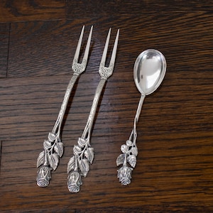 May include: Three silver serving utensils: two forks and a spoon. The forks have two tines and ornate handles with a floral rose and leaf design. The spoon has a rounded bowl and a similar floral handle. All are displayed on a dark wood surface.