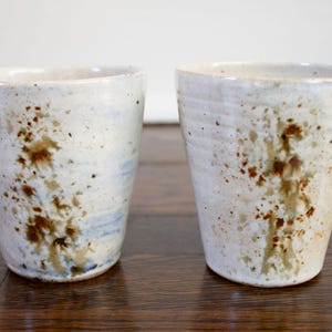 May include: Two handmade ceramic cups. The cups are white with brown and blue speckled patterns. The cups have a slightly tapered shape and a smooth, unglazed rim. The cups are likely used for drinking beverages.