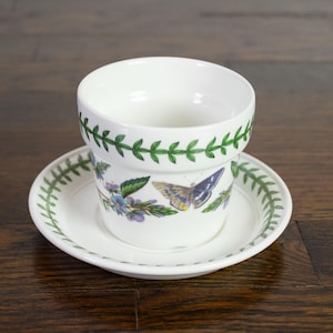 May include: A white ceramic flower pot with a matching saucer, featuring a green leaf vine border around the rim and base. The exterior of the pot is decorated with colourful illustrations of butterflies and purple flowers on a white background.