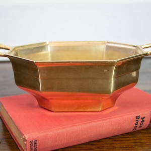 May include: A polished brass bowl with an octagonal shape and two side handles, featuring a smooth metallic finish. The bowl is resting on a red hardcover book with visible text on the spine that reads Collins Dr. Kildare Max Brand.