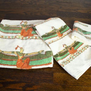 Puede incluir: Un juego de tres toallas de color blanco roto con un patrón repetitivo de una figura de estilo egipcio con alas extendidas en tonos naranja, verde y negro. Las toallas se muestran en varios tamaños, incluyendo una toalla de baño grande y toallas de mano.