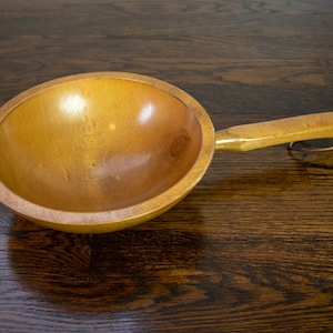May include: A wooden bowl with a long handle and a copper wire loop. The bowl is a light brown color and appears to be made of a single piece of wood. The handle is attached to the side of the bowl.