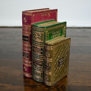 May include: Three decorative book sculptures in varying sizes. The largest is burgundy with gold accents, the middle is green with gold, and the smallest is gold. Each has embossed details and text.