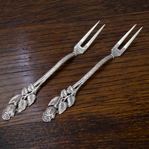 May include: Two silver serving forks with ornate floral and leaf detailing. The tines are slender and the handles feature a rose design at the base. The forks are set against a dark wood grain background.