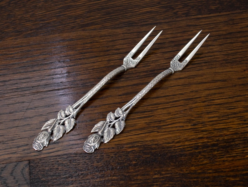 May include: Two silver serving forks with ornate floral and leaf detailing. The tines are slender and the handles feature a rose design at the base. The forks are set against a dark wood grain background.