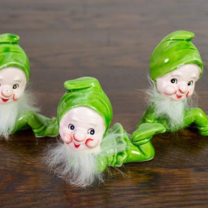 Könnte beinhalten: Drei Keramik-Elf-Figuren mit grünen Hüten und Outfits, weißen Bärten und rosigen Wangen. Die Elfen befinden sich in einer kriechenden Position, mit ausgestreckten Beinen. Die Figuren stehen auf einer dunklen Holzoberfläche.