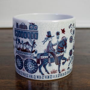 Könnte beinhalten: Zylindrischer Behälter aus weißer Keramik mit einer blau-weißen Illustration eines Pferdewagens und Reitern. Das Design umfasst Blumen- und Vogelmotive mit einem Craquelé-Finish.