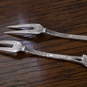 May include: Two silver serving forks with ornate, scrolled designs on the tines and handles. The handles have a textured, branch-like appearance with leaf details. The forks are set against a dark wood background.