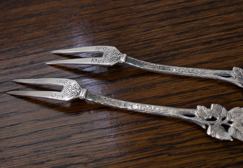May include: Two silver serving forks with ornate, scrolled designs on the tines and handles. The handles have a textured, branch-like appearance with leaf details. The forks are set against a dark wood background.