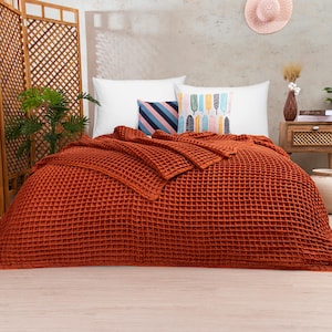 Peut inclure: Une couverture en tissu gaufré de couleur rouille sur un lit avec des oreillers blancs et à motifs.